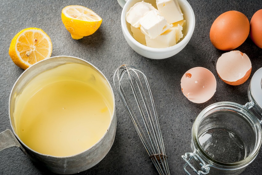Traditionelle Basissaucen. Französische Küche. Sauce Hollandaise in einem Metalltopf mit Zutaten zum Kochen - Eier, Butter, Zitronen. Auf einem schwarzen Steintisch. Draufsicht des Kopierbereichs - Oliver's Sauce Hollandaise