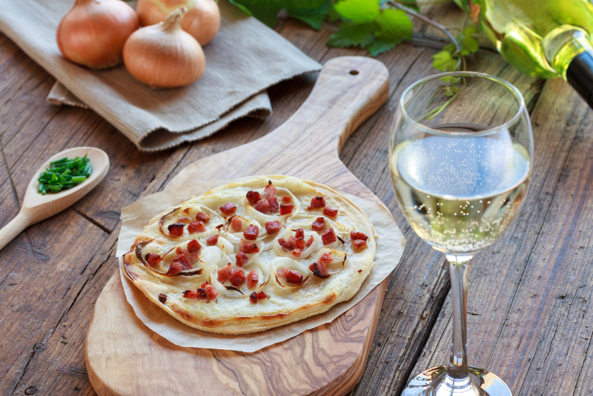 Ein kleiner Flammkuchen ist auf einem Brett drapiert, daneben steht ein Glas Wein
