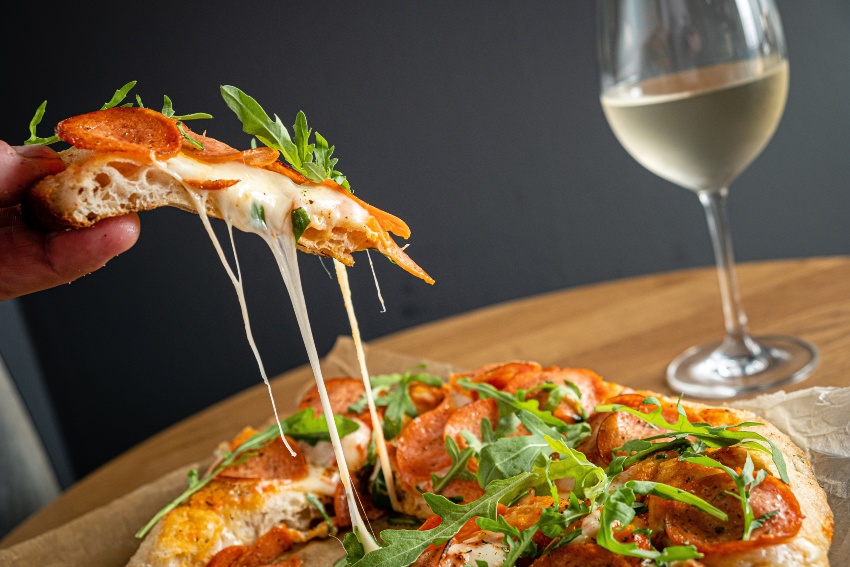 Jemand nimmt sich ein Stueck Pizza, daneben steht ein Glas Weisswein